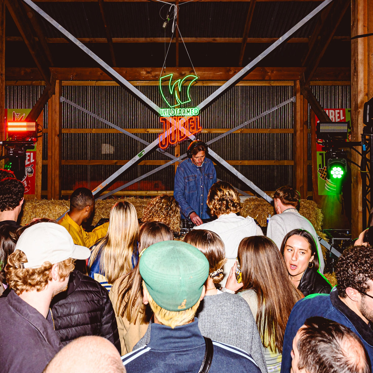 Crowd of people at a Jubel x Wildfarmed party barn-style venue with neon signs, stage lights and Andy Cato DJing.